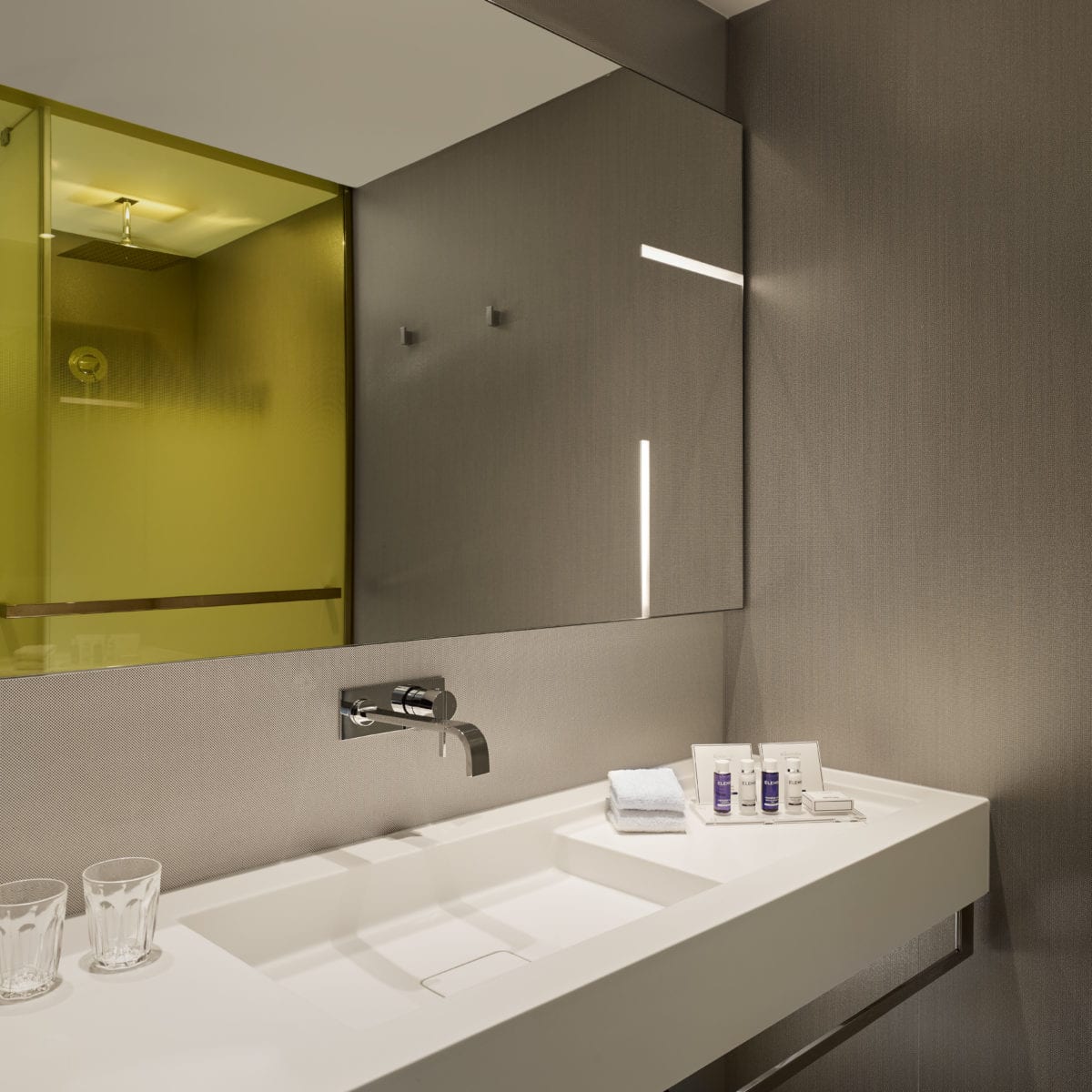 A shot of the bathroom with the yellow shower reflected in the mirror. an assortment of blue and white skincare creams and two glasses in view.