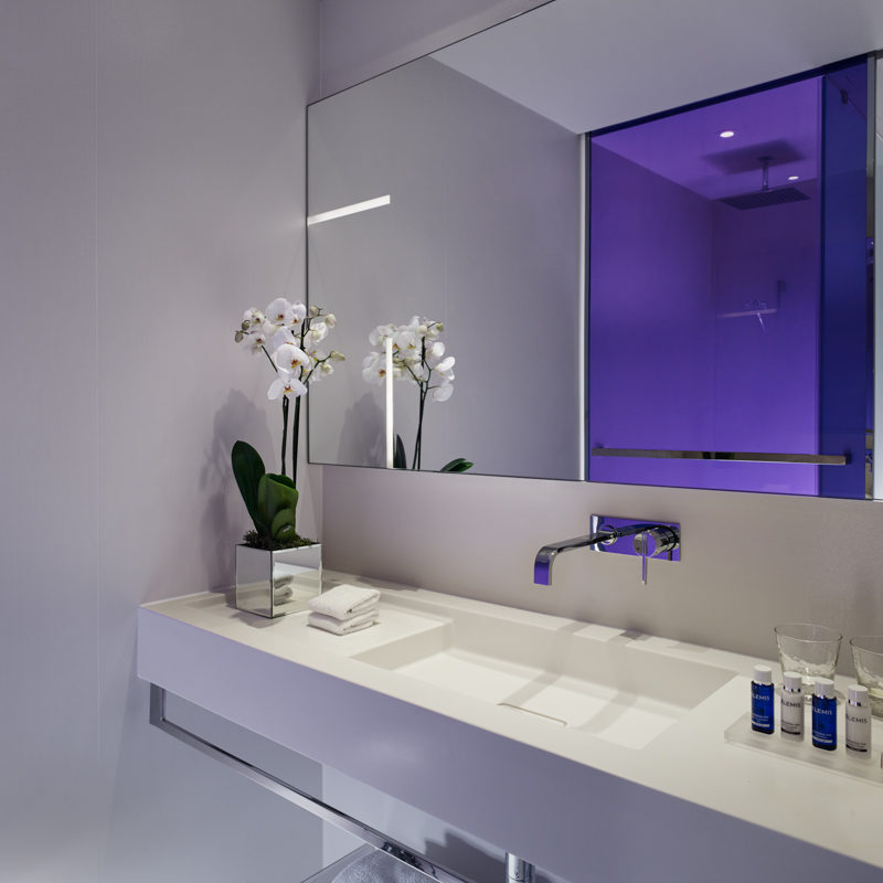 A bright white bathroom with a tall flower on the side and a blue shower screen door reflected in the mirror