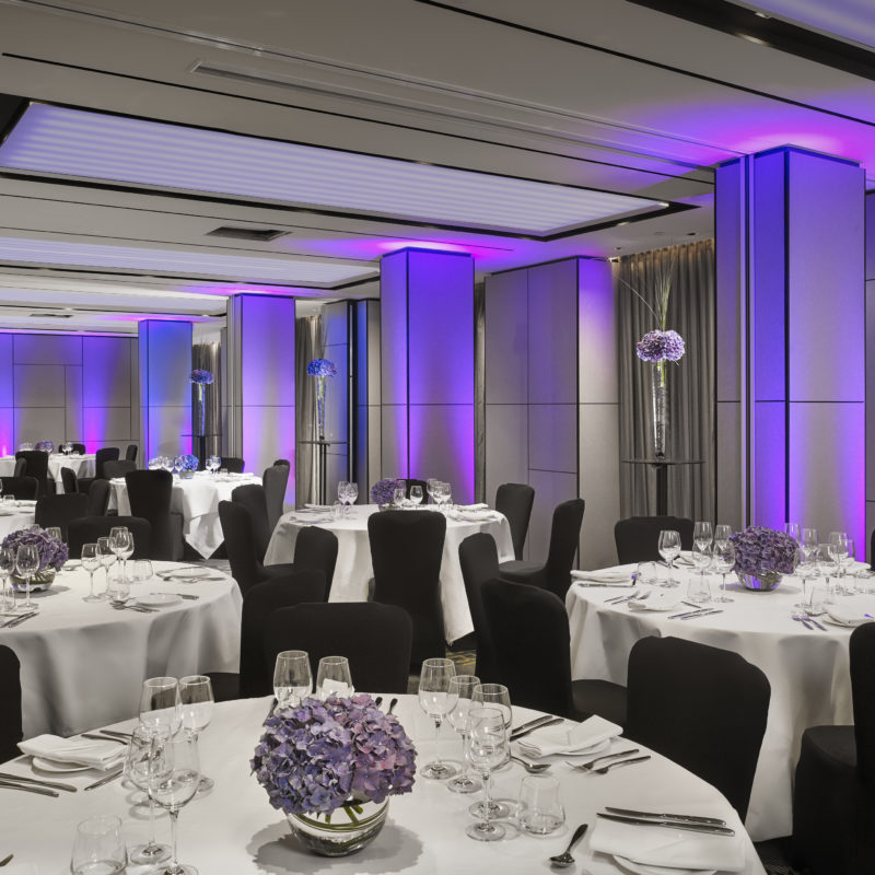 A room full of circular tables with white tablecloths over them and purple flowers decorating them