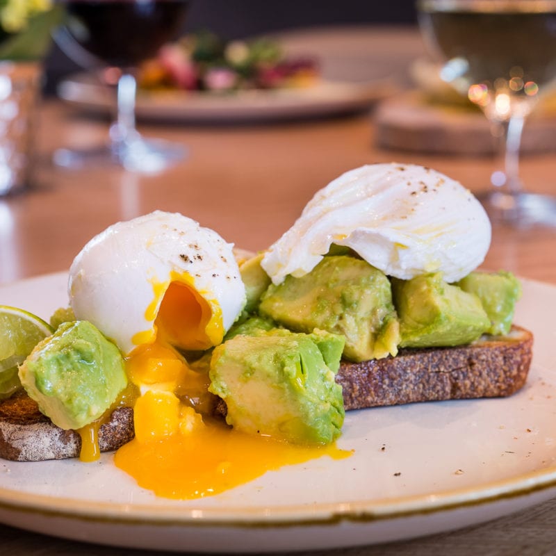 Two pieces of toasted topped with avocado with a poached egg on both. Yellow yolk oozing out of one egg