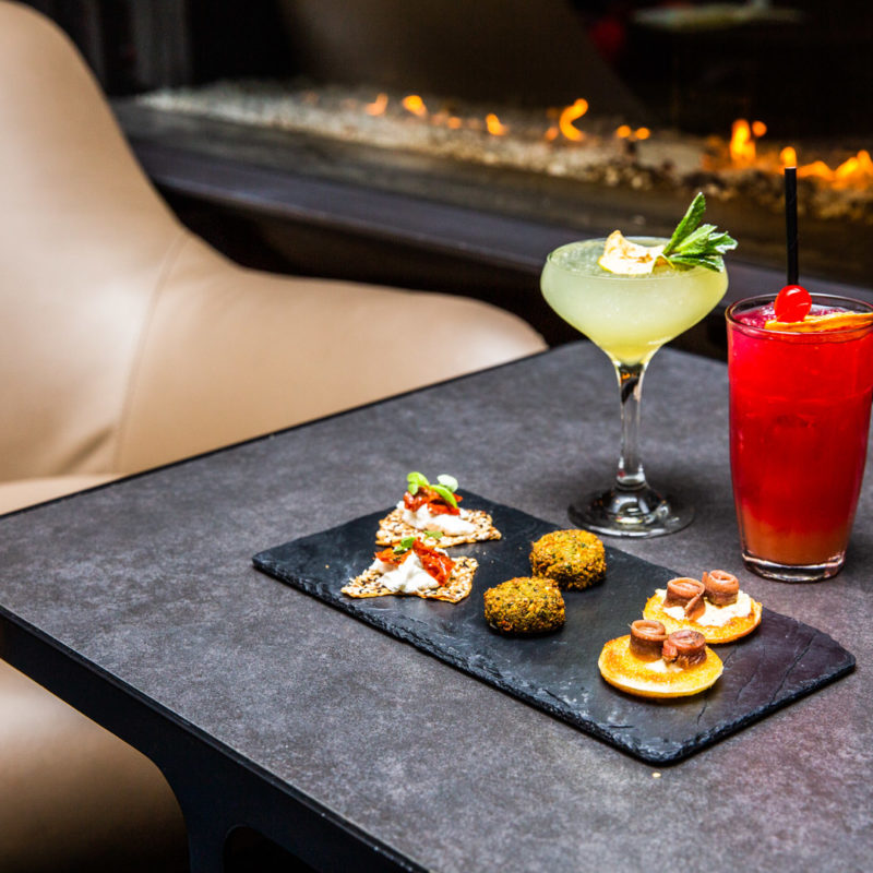 A small tray of aperitivo in front of two brightly coloured cocktails one bright red and one white and yellow