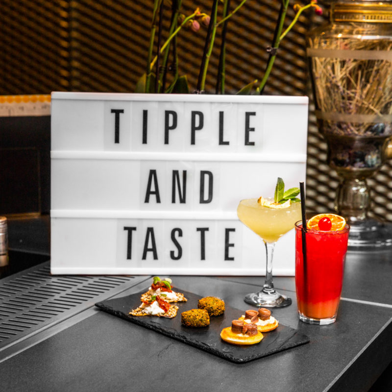 White Sign with the words TIPPLE AND TASTE with a small tray of appetizers in front and two cocktails