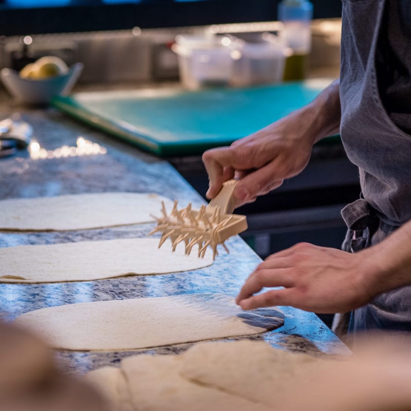 A Florentine chef using a wooden kitchen utensil to on flatbread dough