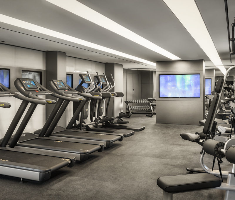 A gym filled with silver gym equipment and blue television screens in front of them