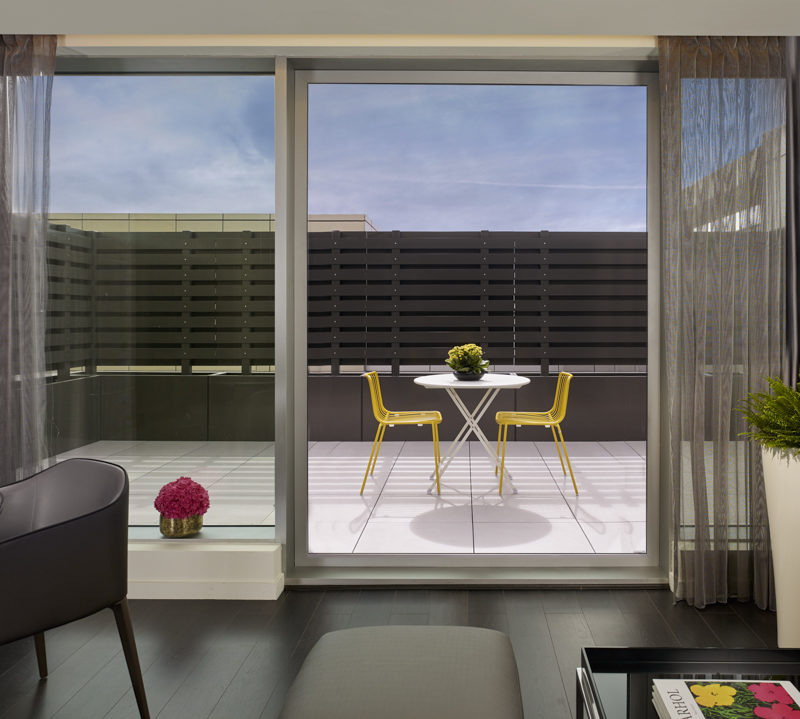 an open screen door leading onto a sunny terrace with a set of white and yellow table and chairs