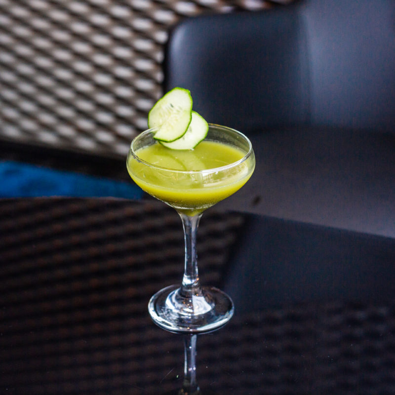 A green cocktail with in a sparkling coupe glass garnished with cucumber