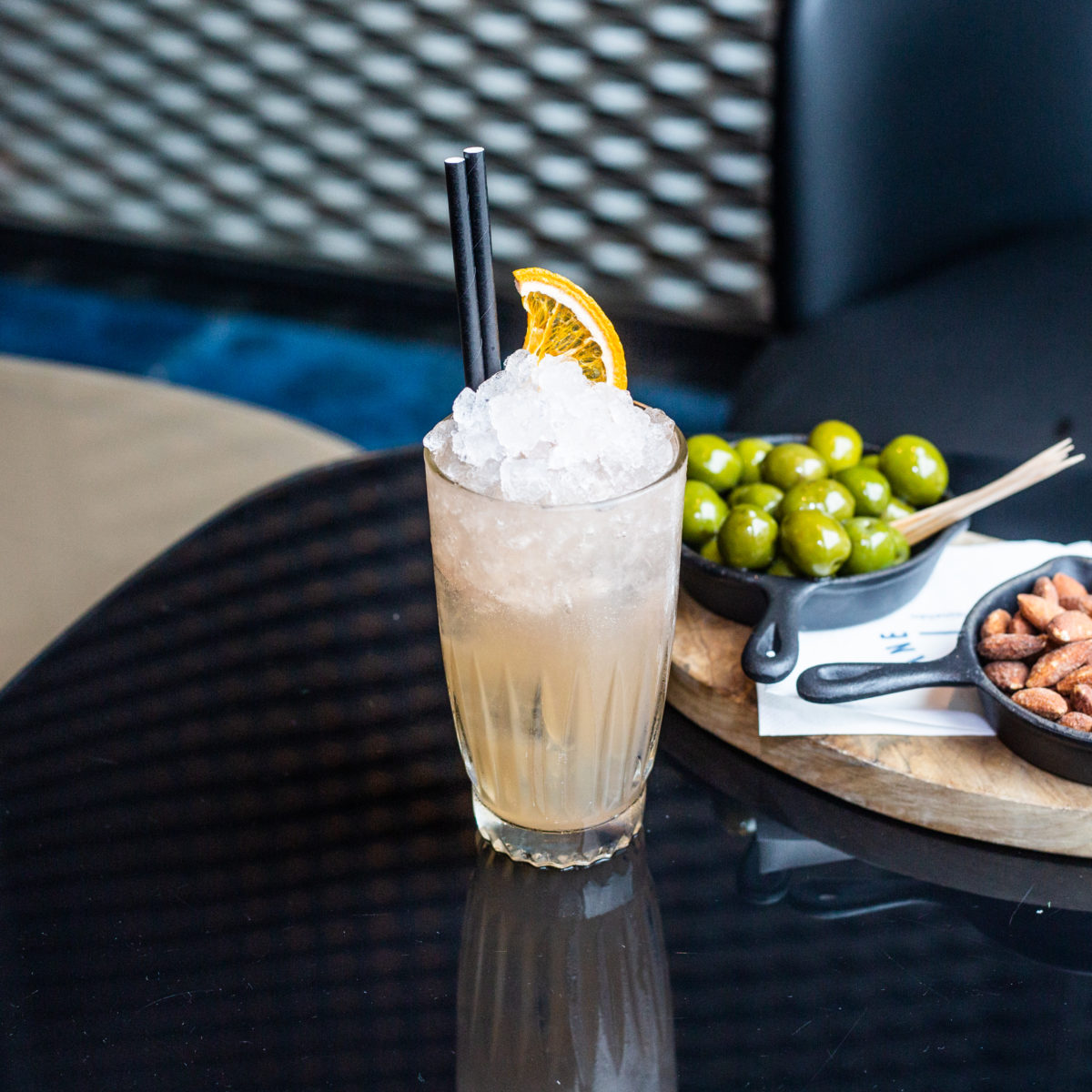 An orange cocktail filled up with ice and garnished with a wedge of orange in front of a plate of green olives and nuts