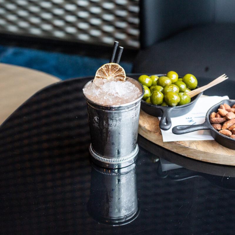 A metal cup with filled with ice and an orange cocktail with a slice of orange on top in front of green olives and nuts