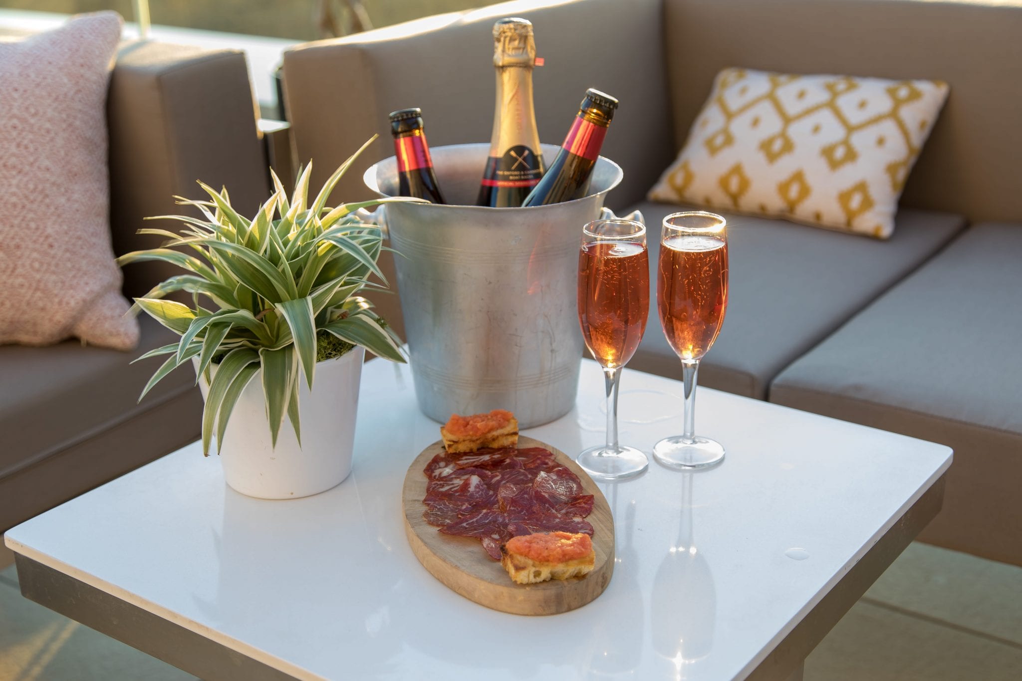 A silver bucket full of gold topped champagne behind a platter of red cured meats and two champagne flutes