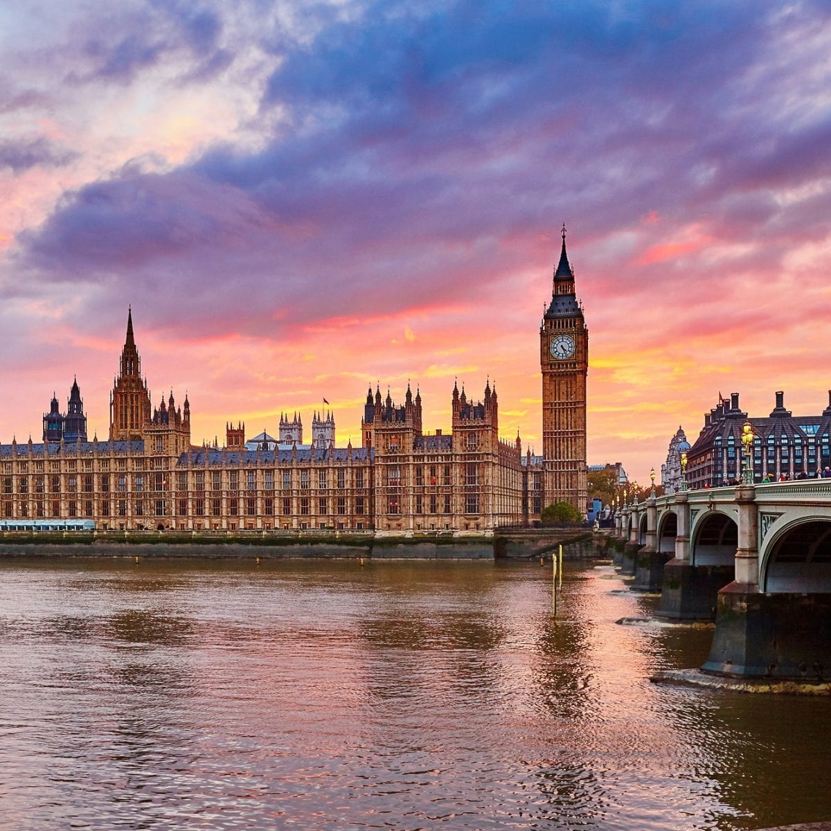The famous Houses of Parliament and Big Ben pictures by the River Thames with a pink and orange sunset behind the iconic London attractions